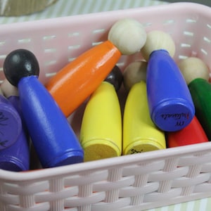 Puede incluir: Una cesta de mimbre rosa llena de figuritas de madera. Las figuritas están pintadas en varios colores, incluyendo azul, naranja, amarillo, verde y rojo. Algunas de las figuritas tienen nombres escritos en ellas, como "Mr. Nathaniel" y "Mr. Nathaniel".
