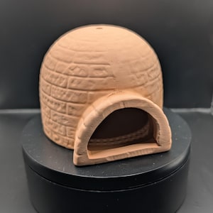 May include: A small, terracotta-coloured ceramic igloo with a rounded dome and a rectangular entrance. The igloo is constructed with a brick-like pattern and sits on a black, circular base. The overall design is simple and rustic.
