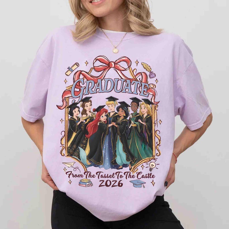 Pode incluir: Camiseta lil&aacute;s claro com um gr&aacute;fico de princesas de desenho animado em trajes de formatura. A palavra "GRADUATE" &eacute; exibida acima das princesas, com o texto "From The Tassel To The Castle 2026" abaixo.