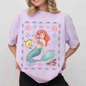 Op de afbeelding: Een lichtpaars t-shirt met korte mouwen en een rechthoekige grafische print van een zeemeermin met rood haar en een groene staart, een gele en blauwe vis en een kleine rode krab. Het ontwerp bevat schelpen, zeesterren en een zonnestralenrand.