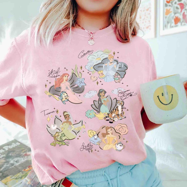 May include: A light pink t-shirt featuring a graphic print of various animated princesses, including Ariel, Cinderella, Jasmine, Tiana, and Belle, accompanied by their respective sidekicks and signatures, with small star and cloud accents throughout.