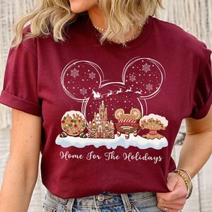 May include: Burgundy t-shirt featuring a festive holiday design. The design includes a Mickey Mouse head outline with snowflakes, a gingerbread castle, and the text "Home for The Holidays".