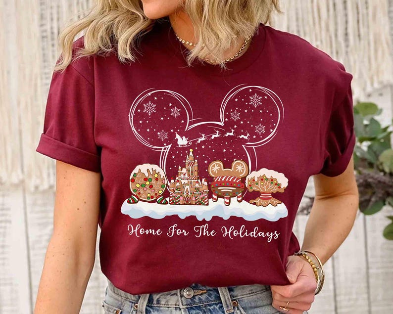 May include: Burgundy t-shirt featuring a festive holiday design. The design includes a Mickey Mouse head outline with snowflakes, a gingerbread castle, and the text "Home for The Holidays".