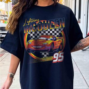 May include: Black t-shirt with a graphic of Lightning McQueen from the film Cars. The design features the text "Lightning McQueen" in yellow and orange, a checkered flag, and the number "95" in white.