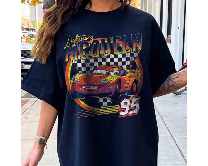 May include: Black t-shirt with a graphic of Lightning McQueen from the film Cars. The design features the text "Lightning McQueen" in yellow and orange, a checkered flag, and the number "95" in white.