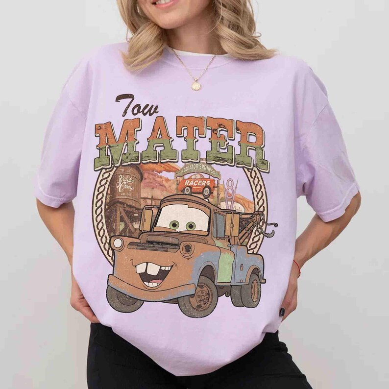 K&ouml;nnte beinhalten: Ein hellviolettes Kurzarm-T-Shirt mit einem Grafikdruck der Figur Tow Mater aus dem Film Cars. Das Design zeigt den Schriftzug Tow Mater, einen braunen Abschleppwagen, einen Wasserturm und eine W&uuml;stenlandschaft innerhalb eines kreisf&ouml;rmigen Rahmens.