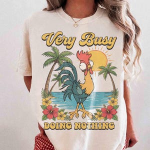 以下が含まれることがあります： クリーム色のTシャツ。熱帯のビーチのシーンと漫画のオンドリが描かれています。シャツには「Very Busy Doing Nothing」という文字が黄色とオレンジ色で書かれています。ヤシの木、花、太陽もデザインの一部です。