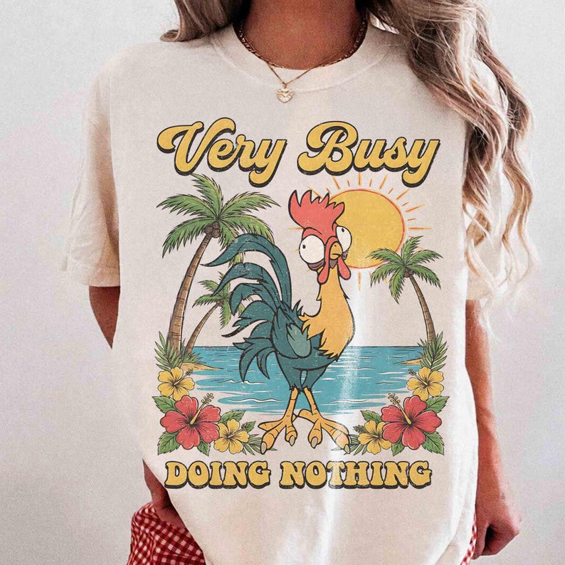 以下が含まれることがあります： クリーム色のTシャツ。熱帯のビーチのシーンと漫画のオンドリが描かれています。シャツには「Very Busy Doing Nothing」という文字が黄色とオレンジ色で書かれています。ヤシの木、花、太陽もデザインの一部です。