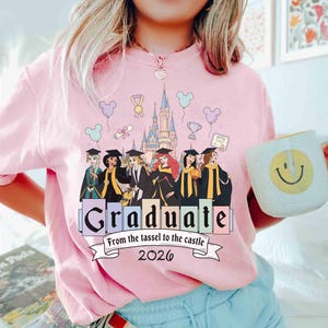 May include: Pink t-shirt with a graduation theme. The shirt features a graphic of Disney princesses in graduation attire, a castle, and the word "Graduate" with the text "From the tassel to the castle 2026". A white mug with a smiley face is held in the image.