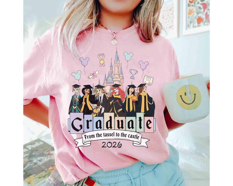 May include: Pink t-shirt with a graduation theme. The shirt features a graphic of Disney princesses in graduation attire, a castle, and the word "Graduate" with the text "From the tassel to the castle 2026". A white mug with a smiley face is held in the image.