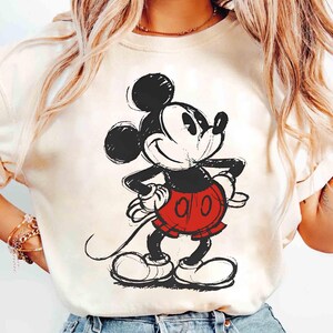 May include: Cream-colored t-shirt featuring a sketch-style illustration of Mickey Mouse. The cartoon character is depicted in black, red, and white, with his iconic shorts and gloves. The design is centered on the shirt.