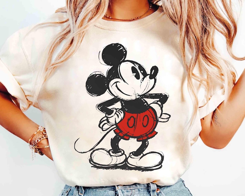 May include: Cream-colored t-shirt featuring a sketch-style illustration of Mickey Mouse. The cartoon character is depicted in black, red, and white, with his iconic shorts and gloves. The design is centered on the shirt.