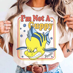 Op de afbeelding: Cr&egrave;mekleurig T-shirt met een cartoonafbeelding van Bot uit De Kleine Zeemeermin, met de tekst "I'm Not A Guppy". Het ontwerp bevat een gele vis met blauwe strepen en een boze uitdrukking. Het woord "Flounder" staat eronder.