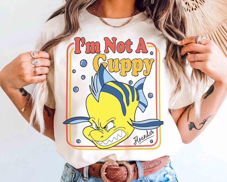 Op de afbeelding: Cr&egrave;mekleurig T-shirt met een cartoonafbeelding van Bot uit De Kleine Zeemeermin, met de tekst "I'm Not A Guppy". Het ontwerp bevat een gele vis met blauwe strepen en een boze uitdrukking. Het woord "Flounder" staat eronder.