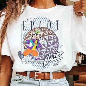 May include: White t-shirt featuring a graphic of Goofy in a colorful outfit, with the text "EPCOT Center" and "Walt Disney World 1982". The design includes a geometric sphere and a retro aesthetic.