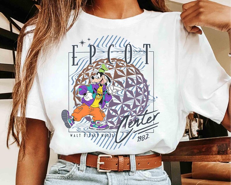 May include: White t-shirt featuring a graphic of Goofy in a colorful outfit, with the text "EPCOT Center" and "Walt Disney World 1982". The design includes a geometric sphere and a retro aesthetic.