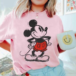 May include: A light pink t-shirt featuring a black and red sketch of Mickey Mouse. The cartoon character is smiling with his hands on his hips. A white mug with a yellow smiley face is held in the hand.