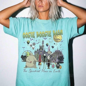 Puede incluir: Una camiseta de color turquesa claro con un estampado gr&aacute;fico que incluye el texto OOGIE BOOGIE BASH y The Spookiest Place on Earth. El dise&ntilde;o muestra varios personajes animados, un castillo, una luna y fuegos artificiales oscuros.