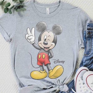 May include: A gray t-shirt featuring a sketch of Mickey Mouse giving a peace sign. Mickey is wearing red shorts and yellow shoes. The Disney logo is visible. The shirt is paired with blue jeans and white sneakers.