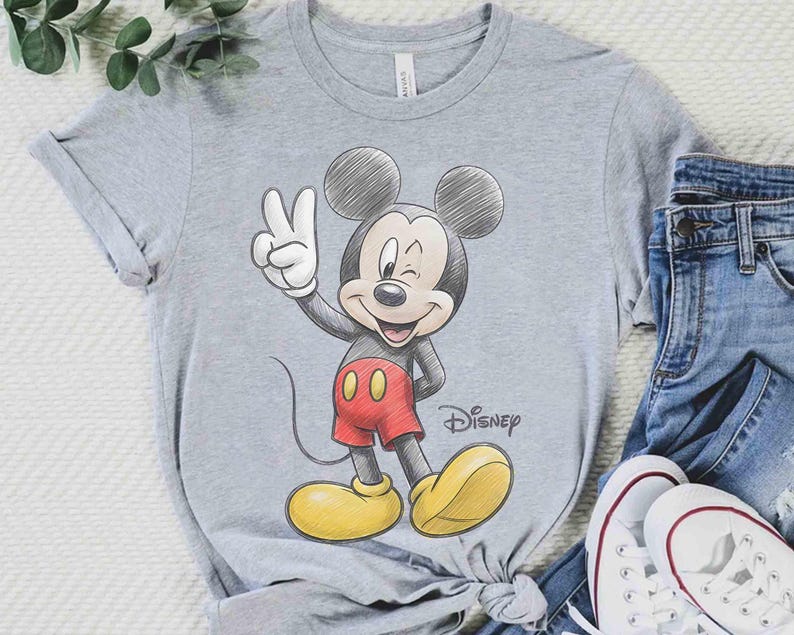 May include: A gray t-shirt featuring a sketch of Mickey Mouse giving a peace sign. Mickey is wearing red shorts and yellow shoes. The Disney logo is visible. The shirt is paired with blue jeans and white sneakers.