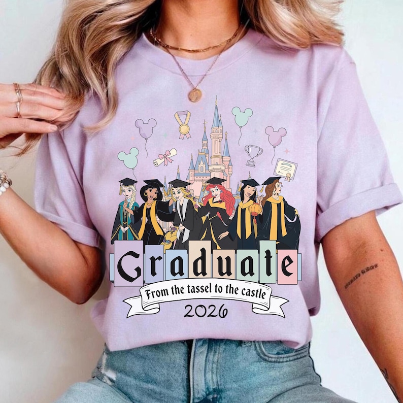 May include: A lilac-colored t-shirt featuring a graphic of Disney princesses in graduation attire in front of a castle. The word "Graduate" is displayed prominently, with the text "From the tassel to the castle 2026" below.