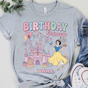 May include: A grey t-shirt with a colourful graphic. It features the words "BIRTHDAY Princess" and the number "5". The design includes a castle, balloons, fireworks, and a cartoon princess in a yellow dress.