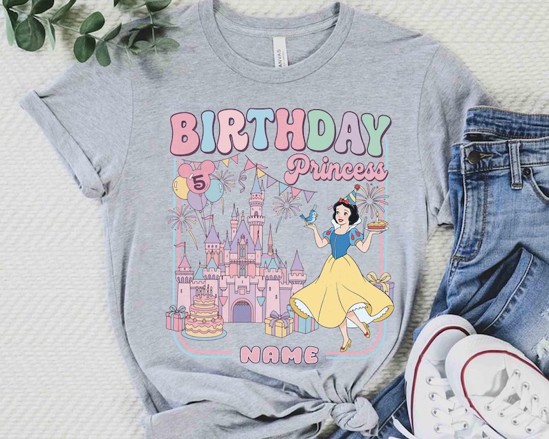 May include: A grey t-shirt with a colourful graphic. It features the words "BIRTHDAY Princess" and the number "5". The design includes a castle, balloons, fireworks, and a cartoon princess in a yellow dress.