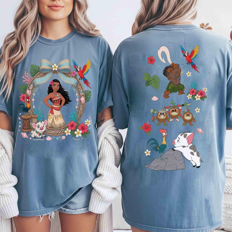 May include: A blue t-shirt featuring a front graphic of Moana framed by a floral wreath with Pua and Heihei, and a back graphic showing Maui, Kakamora, Heihei, and Pua surrounded by tropical flowers, leaves, and seashells in vibrant colors.