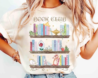 Puede incluir: Camiseta color crema con una ilustración caprichosa de tres estanterías llenas de libros coloridos y objetos temáticos de cuentos de hadas. El texto "BOOK CLUB" está arqueado sobre las estanterías. El diseño incluye una rana, una rosa en una cúpula de cristal y un dragón.