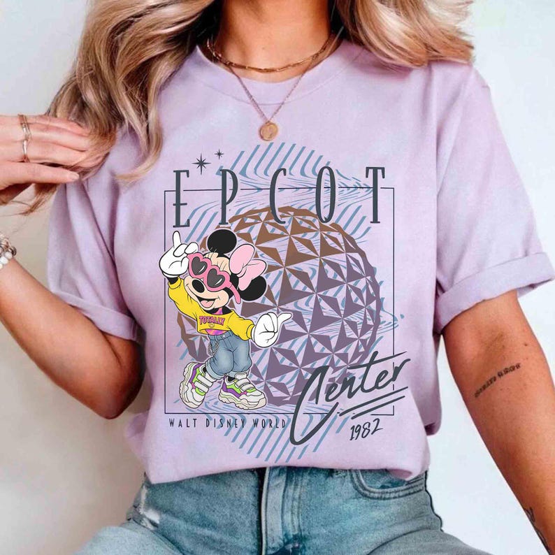May include: A lilac-colored t-shirt featuring a graphic of Minnie Mouse wearing sunglasses and a yellow jacket, with the text "EPCOT Center 1982" and a geometric sphere design. The shirt is part of a Walt Disney World collection.