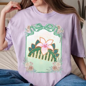Puede incluir: Una camiseta lila con un dise&ntilde;o floral que presenta un peine, un lirio y un lazo. El dise&ntilde;o est&aacute; enmarcado por un borde verde y rodeado de flores. La camiseta est&aacute; hecha de un material suave.