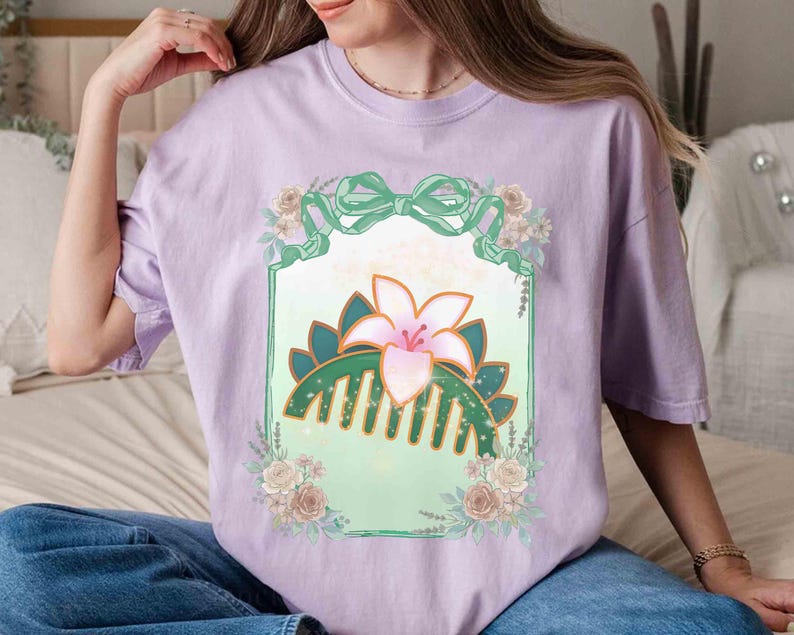 Puede incluir: Una camiseta lila con un dise&ntilde;o floral que presenta un peine, un lirio y un lazo. El dise&ntilde;o est&aacute; enmarcado por un borde verde y rodeado de flores. La camiseta est&aacute; hecha de un material suave.