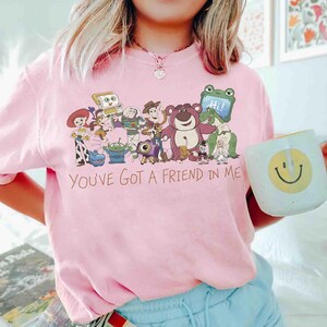 Op de afbeelding: Roze crewneck T-shirt met een afbeelding van Toy Story-personages en de tekst "You've Got a Friend in Me". Een witte mok met een gele smiley wordt in de hand gehouden. Het shirt heeft een lichtroze kleur.