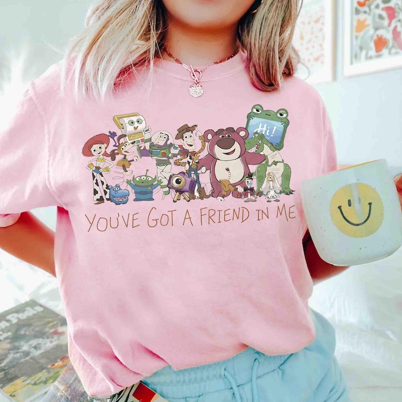 Op de afbeelding: Roze crewneck T-shirt met een afbeelding van Toy Story-personages en de tekst "You've Got a Friend in Me". Een witte mok met een gele smiley wordt in de hand gehouden. Het shirt heeft een lichtroze kleur.