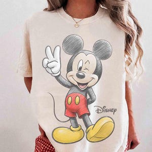 May include: Cream-colored t-shirt featuring a sketch-style illustration of Mickey Mouse winking and giving a peace sign. Mickey is wearing red shorts and yellow shoes. The Disney logo is printed on the shirt.