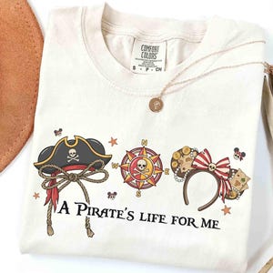 Könnte beinhalten: Cremefarbenes T-Shirt mit Piratenmotiven. Das Design umfasst einen Piratenhut, einen Kompass und Micky-Maus-Ohren mit Schleife, alle mit Totenköpfen und Münzen verziert. Der Text "A Pirate's Life for Me" ist darunter gedruckt.
