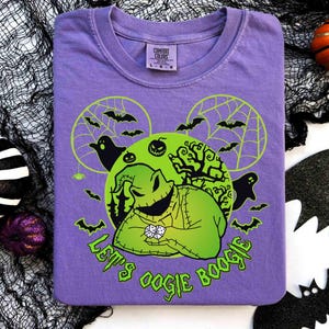 May include: A purple t-shirt featuring a green graphic of a character with dice, bats, and the text "Let's Oogie Boogie". The design includes a green circle with a spiderweb and bats. The shirt is folded on a white surface.