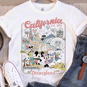 Puede incluir: Camiseta blanca con un gr&aacute;fico colorido de Disneyland que presenta a Mickey Mouse, Minnie Mouse, Donald Duck y otros personajes. El gr&aacute;fico incluye el texto "California Disneyland Est. 1955". La camiseta se combina con vaqueros azules y zapatillas blancas.