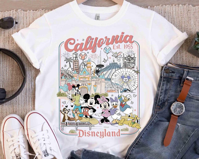 Puede incluir: Camiseta blanca con un gr&aacute;fico colorido de Disneyland que presenta a Mickey Mouse, Minnie Mouse, Donald Duck y otros personajes. El gr&aacute;fico incluye el texto "California Disneyland Est. 1955". La camiseta se combina con vaqueros azules y zapatillas blancas.