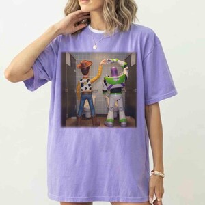 May include: A light purple short-sleeve t-shirt featuring a graphic print of the characters Woody and Buzz Lightyear standing in bathroom stalls, forming a heart shape with their hands above their heads against a tiled wall background.