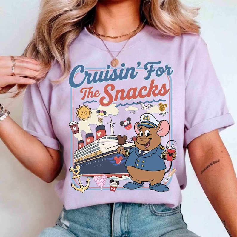 Pode incluir: T-shirt lil&aacute;s claro com o texto "Cruisin' For The Snacks" em azul e laranja. O design apresenta um rato de desenho animado com uniforme de capit&atilde;o segurando um sorvete do Mickey Mouse e uma bebida, com um navio de cruzeiro ao fundo.