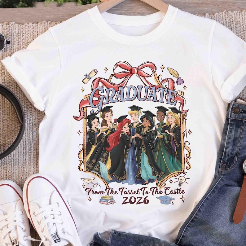 Pode incluir: T-shirt branco com um design de formatura. O design inclui a palavra "GRADUATE" em uma fita vermelha e ilustra&ccedil;&otilde;es de personagens de desenhos animados em trajes de formatura. O texto diz "From The Tassel To The Castle 2026".