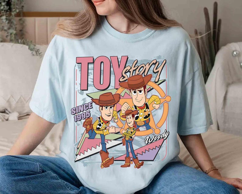 May include: Pale blue t-shirt with a graphic of Woody from Toy Story. The design includes the text "TOY STORY" and "SINCE 1995". Woody is shown in various poses, wearing a cowboy hat and boots. The t-shirt has a loose fit.
