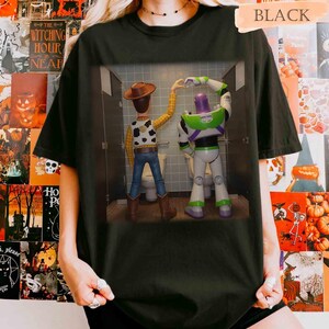May include: A black t-shirt featuring a graphic print of Woody and Buzz Lightyear standing in bathroom stalls, with their arms raised to form a heart shape together. The characters are viewed from behind, showcasing their iconic outfits and colors.