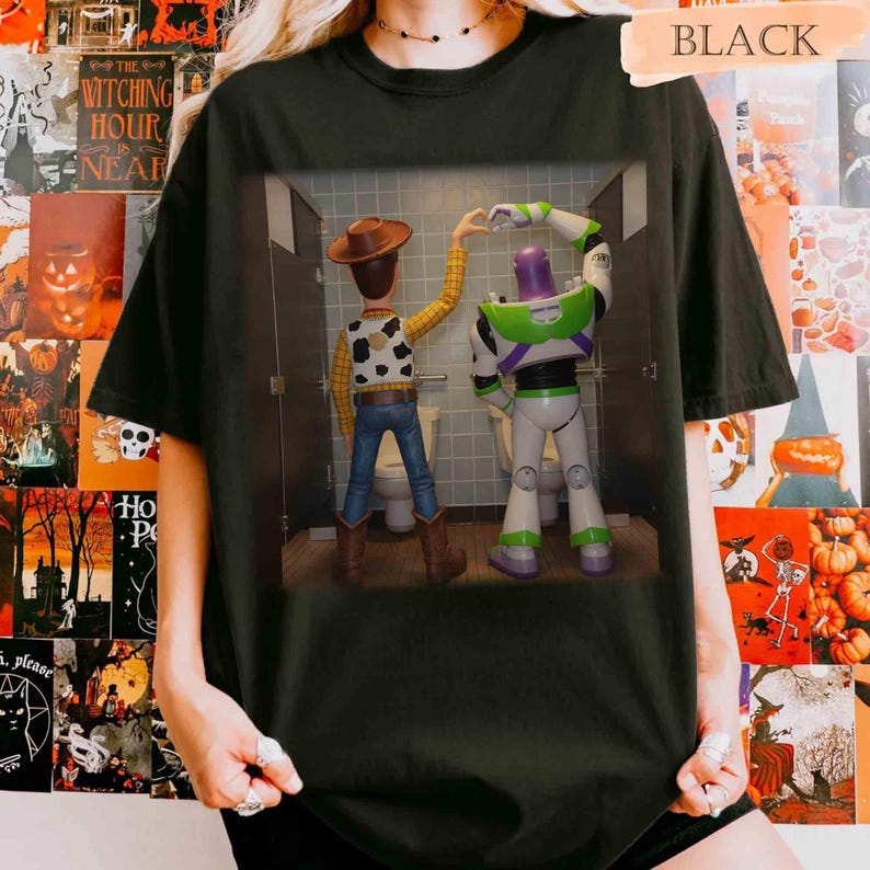 May include: A black t-shirt featuring a graphic print of Woody and Buzz Lightyear standing in bathroom stalls, with their arms raised to form a heart shape together. The characters are viewed from behind, showcasing their iconic outfits and colors.