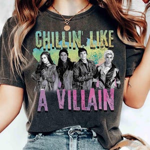 May include: Dark gray t-shirt featuring the phrase "CHILLIN LIKE A VILLAIN" in colorful text above a graphic of four people. The shirt is a comfortable, casual style.
