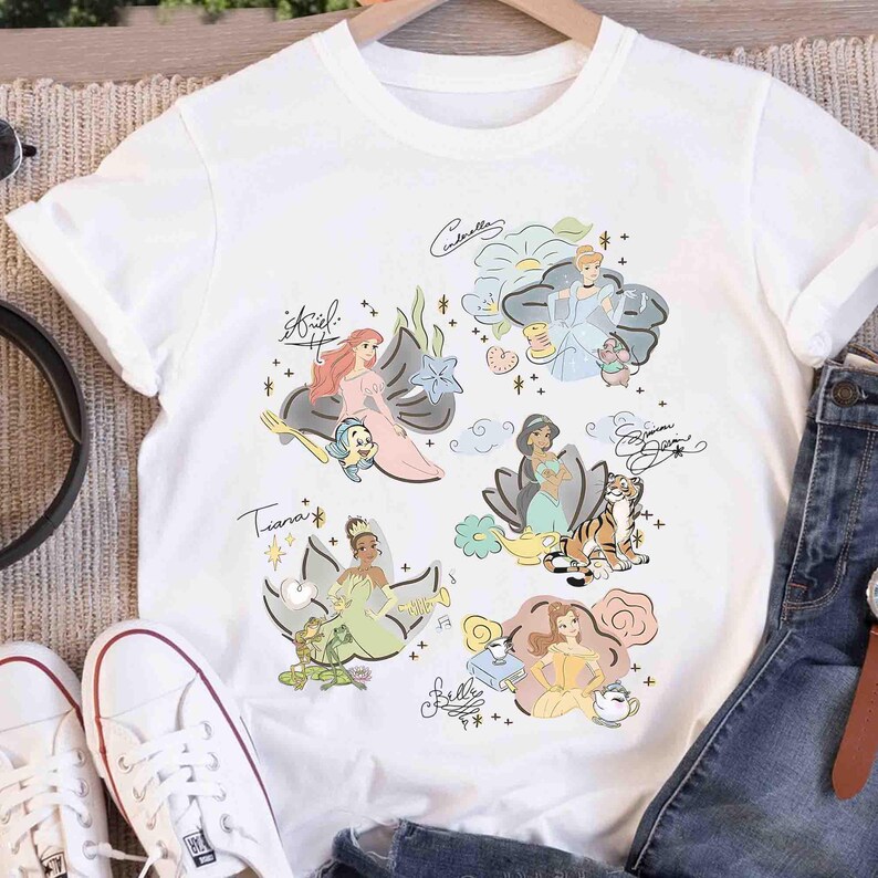May include: A white short-sleeved t-shirt featuring a graphic print of six animated princesses including Ariel, Cinderella, Jasmine, Tiana, and Belle. Each character is accompanied by their signature, a small companion, and decorative floral elements.