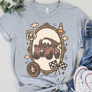 May include: A light gray t-shirt featuring a cartoon image of a brown tow truck inside a decorative frame. The frame includes a Route 66 sign, clouds, a traffic cone, a tire, and checkered flags. The shirt is paired with blue jeans and white sneakers.