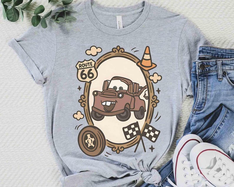 May include: A light gray t-shirt featuring a cartoon image of a brown tow truck inside a decorative frame. The frame includes a Route 66 sign, clouds, a traffic cone, a tire, and checkered flags. The shirt is paired with blue jeans and white sneakers.