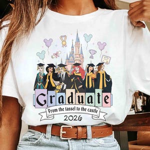 May include: White t-shirt featuring a colorful graphic of Disney princesses in graduation attire in front of a castle. The shirt includes the text "Graduate" and "From the tassel to the castle 2026".